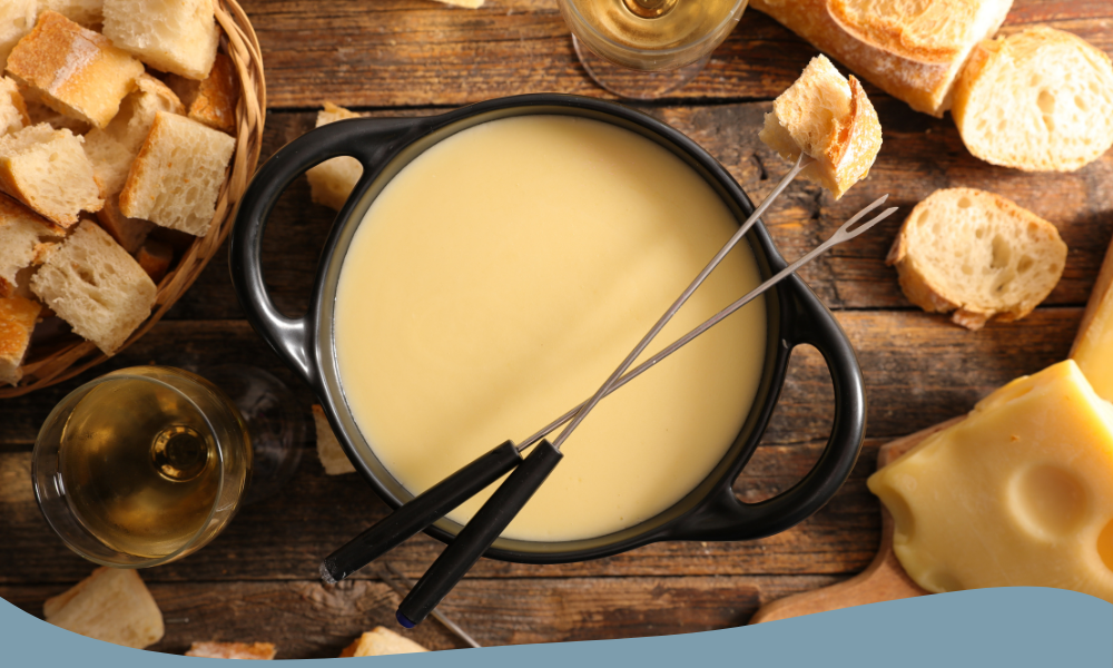 Fondueset placerat på ett rustikt bord, omgiven av brödbitar och ostskivor. Fonduegrytan är fylld med smält ost, och två fonduegafflar håller brödbitar redo att doppas. Ett glas vitt vin kompletterar måltiden.