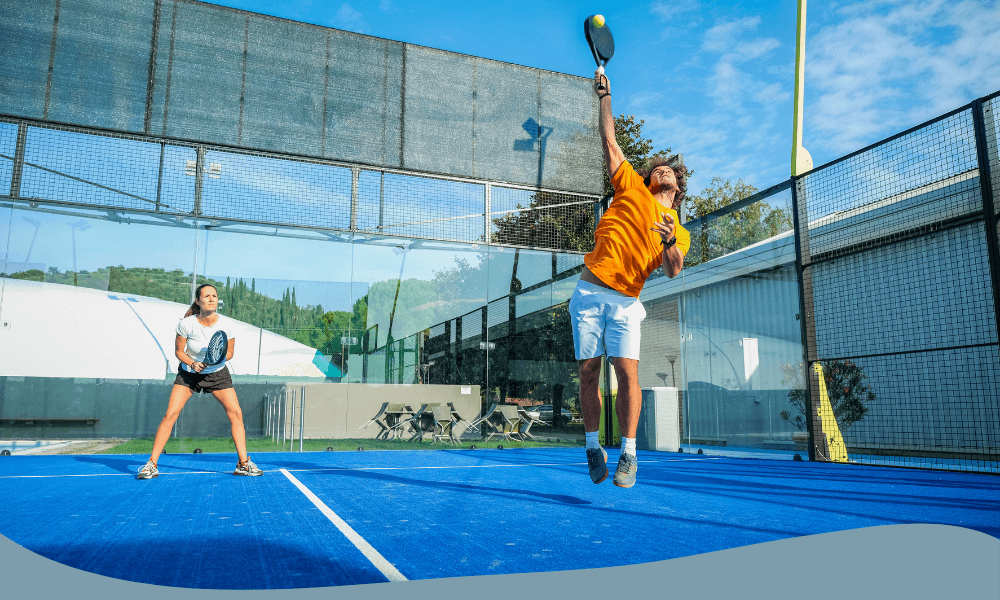 Man och kvinna spelar padel, mannen hoppar och smashar bollen medan kvinnan förbereder sig i försvarsposition.