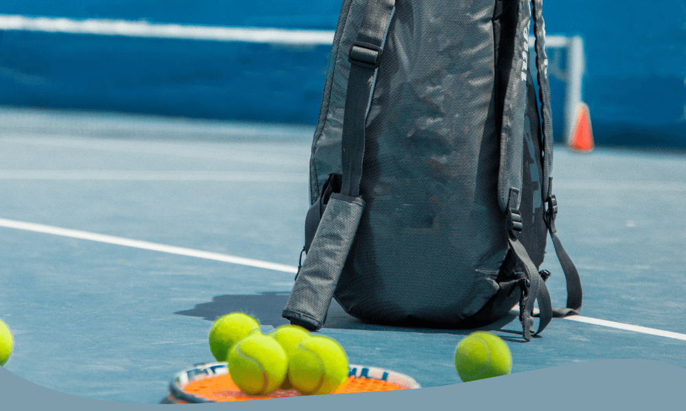 Svart tennisryggsäck placerad bredvid tennisbollar och racket på tennisbana, redo för spel.